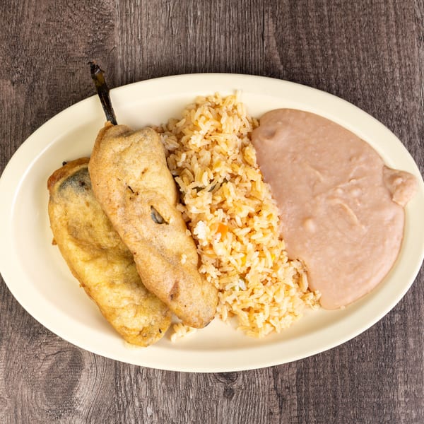 Two chile rellenos, rice, and refried beans on an oval plate on a wooden table.