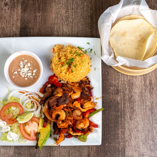 A fajita platter with shrimp, steak, chicken, rice, beans, salad, and tortillas on a wooden table.