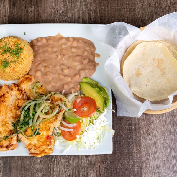 Grilled chicken with rice, refried beans, salad, avocado, and tortillas on a wooden table.