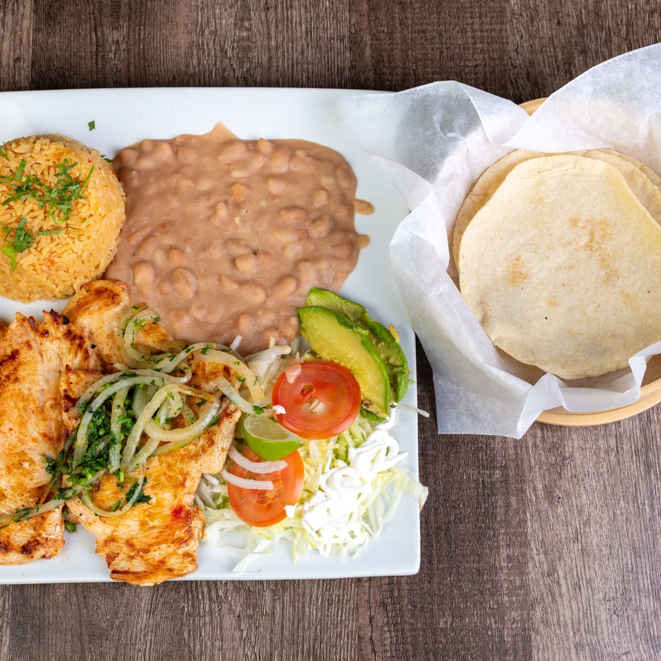 Grilled chicken with rice, refried beans, salad, avocado, and tortillas on a wooden table.