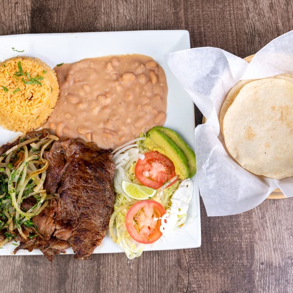 Grilled steak with onions, rice, refried beans, salad, avocado, and tortillas on a wooden table.